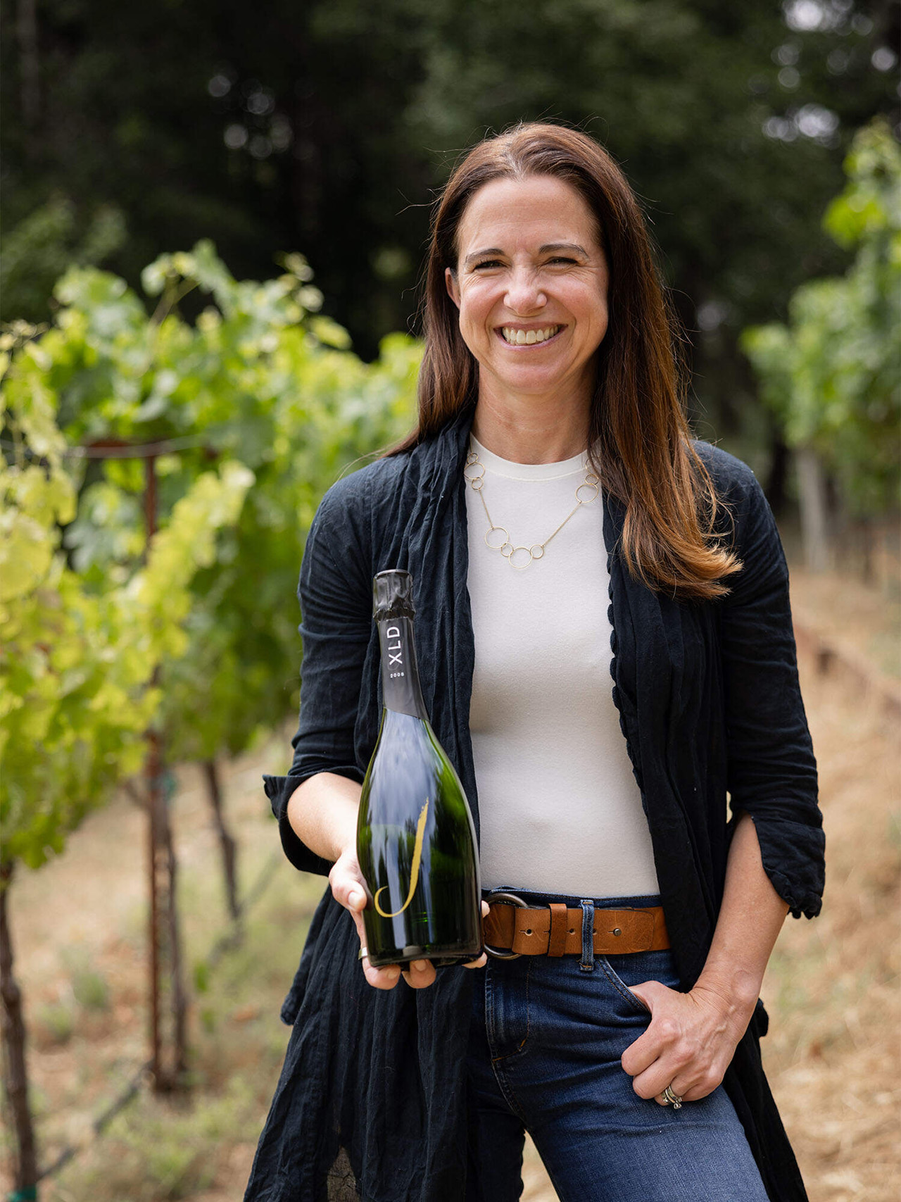 Winemaker, Nicole Hitchcock, standing in a vineyard holding a bottle of J Wine.